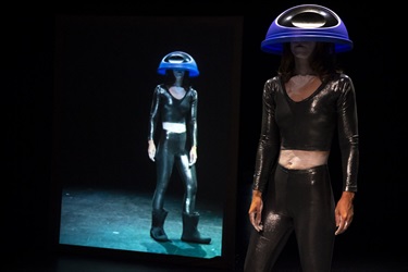 A person wearing a dark body suit wears a large bowl-like helmet on their head. The helmet covers their face. Behind them is a mirrored image.