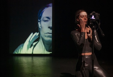 A woman wearing a tight dark body suit stands behind a camera and smears her left cheek. Behind her left shoulder is a projected image of a woman's face.