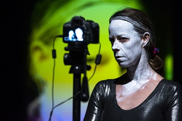 A woman wearing white makeup on her face and neck and a hair band on her head stands in front of a camera and a projection screen.