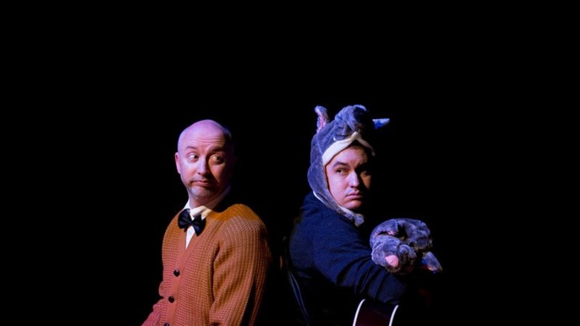Two people sit back to back of one another. The person on the left wears a mustard cardigan and a black bow tie. The person on the right weas a possum costume.