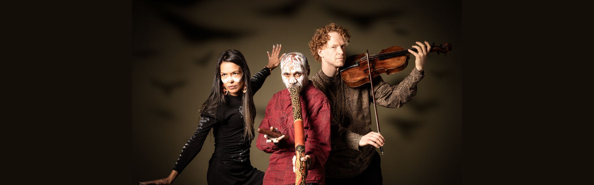 Three people stand side by side: one plays the violin, one plays the didgeridoo and the third holds her arms out to the sides like wings.