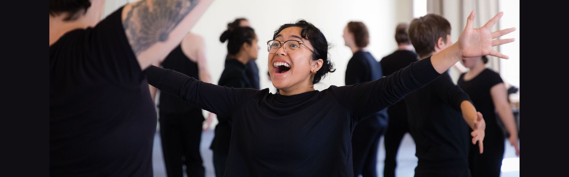 A person with glasses and dark short hair extends their arm out wide with a look of joy and surprise on their face.