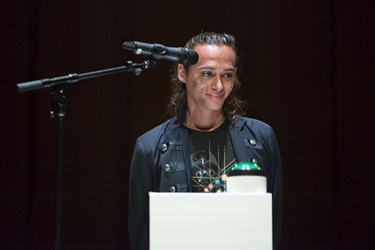 A person with dark hair wears a dark top and stands behind a white podium and a microphone.