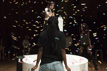 A person wearing jeans and a black top has their back to the camera. Confetti is falling from the ceiling. A white circular platform in in front of the person.