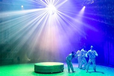 A round platform sit at the centre of the stage. Four people wearing silver outfits stand on the right side of the platform in different robotic poses. A bright light from above the platform shines down.