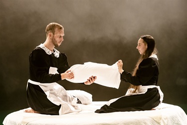 A man with light hair and a woman with long dark hair sit on their legs facing each other. They both wear long black outfits with white waist aprons. They hold a white pillow between them.