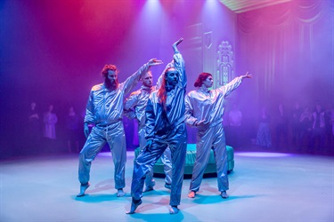 Four people wearing silver track suits cluster together at centre stage. The woman in front stands with one arm in the air and her left foot pointed but resting on the floor out to the side.