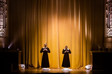 Two people stand with their heads bowed and their hands folded in front of their chests. The wear long black gowns with white collars. They stand in front of a yellow theatre curtain.