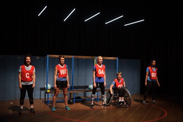 Five women of various ethnicities and abilities wear netball uniforms and stand side by side across a stage.