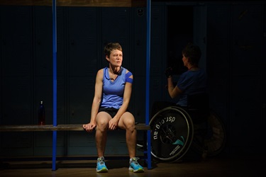 A woman with short dark hairs wears a blue athletic tank top and sits on a gym bench. In front of her is a person in a wheelchair.