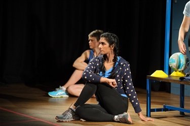 A woman with long dark hair pulled back in a ponytail wears dark pants and a polka dot jumper. She is sitting on the floor with one leg crossed over the other and an arm resting on the knee.
