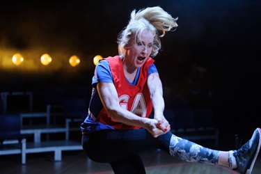A woman with long light hair pulled back in a ponytail wears a red loose tank top and dark pants. She kicks on leg in the air as if punting a ball.
