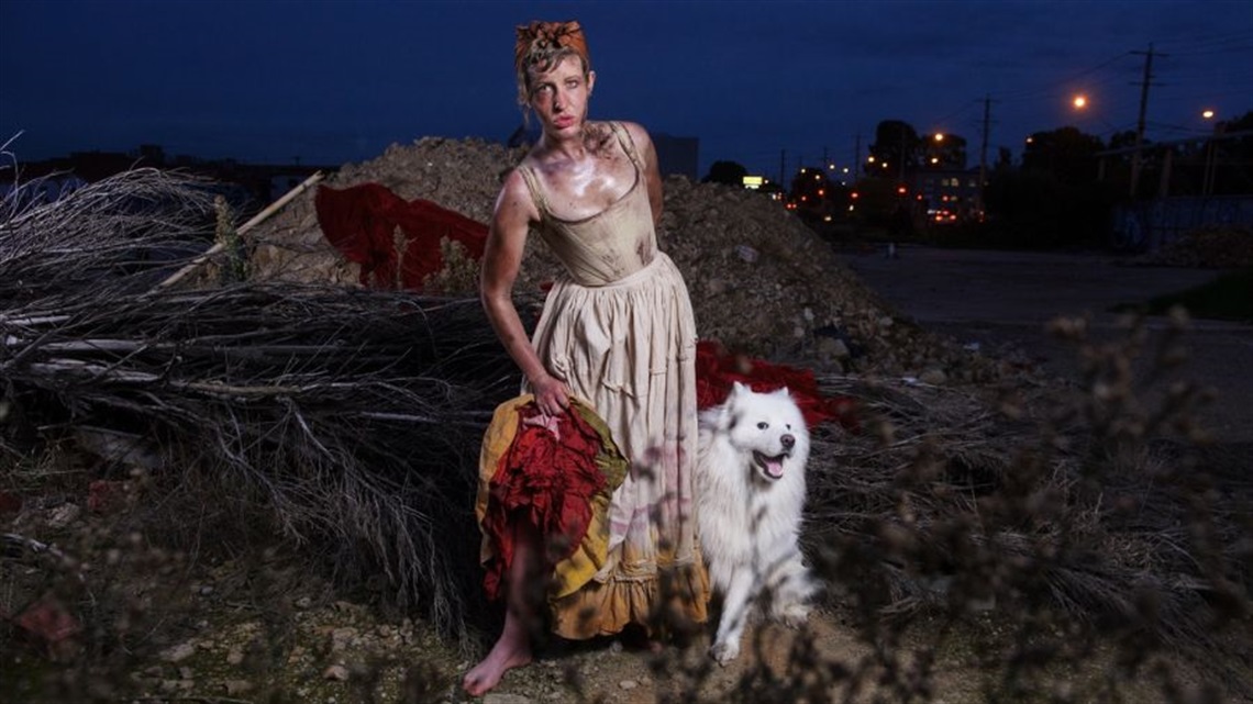 A person wearing a white dress lifts the bottom of the skirt to reveal red and yellow ruffles from underneath. They lean forward and stand with a white dog next to them. Behind is a pile of dirt and a large tree branch.