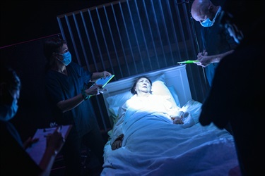 A woman with short dark hair is lying in a hospital bed while three other people wearing medical masks surround her. A blue light shines on the woman in the bed.