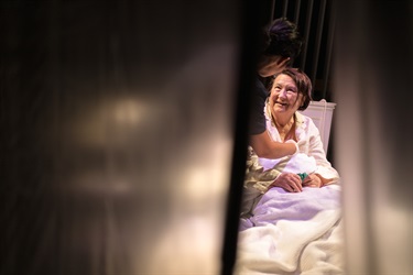 Through a slight opening of a blurring clear curtain, a woman with short dark hair sits up in a bed. Another woman with long dark hair leans over her checking her heart.