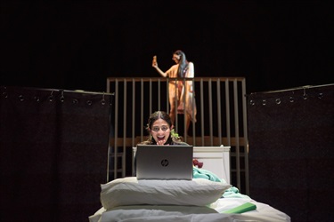 A woman sits on a bed behind an Apple computer laptop screen. Another woman is behind her on a raised platform. She holds a mobile phone as if video chatting.