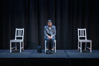 A woman with long dark hair sits on a chair in the centre of a black stage. On either side of her are empty white chairs.