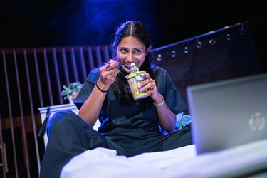 A woman with dark hair sits on a white bad wearing dark scrubs. She holds a can of Chinese noodles and a fork to her mouth.