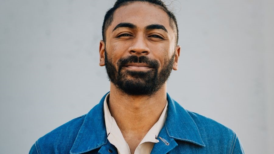 A man with short dark hair and a beard wears a blue denim jacket and a white shirt underneath. He smiles tot he camera.