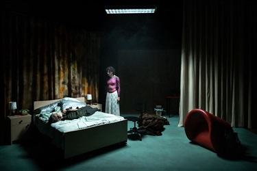 A dark setting with one person laying on their back on a bed and another person wearing a red shirt and white skirt standing over them.