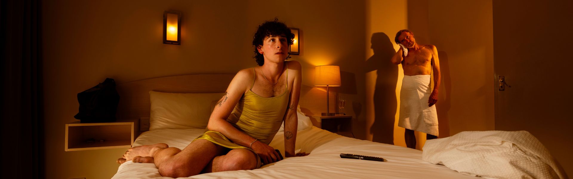 A person wearing a gold dress sits on a hotel bed looking outwards. Another person is in the background wearing a towel around their waist looking at the person on the bed.