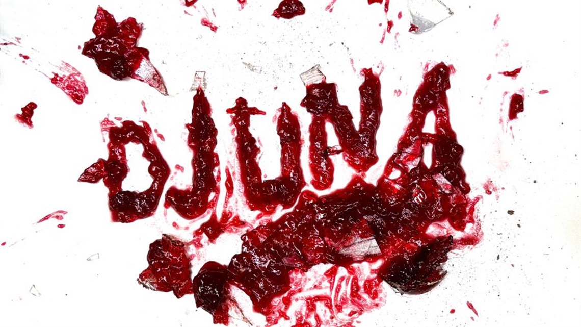 The word Djuna written out in red thick jam with shattered glass pieces scattered on a white backdrop.