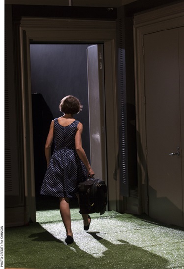 A woman with short dark hair wears a sleeves dress and is walking out a doorframe carrying a suitcase.