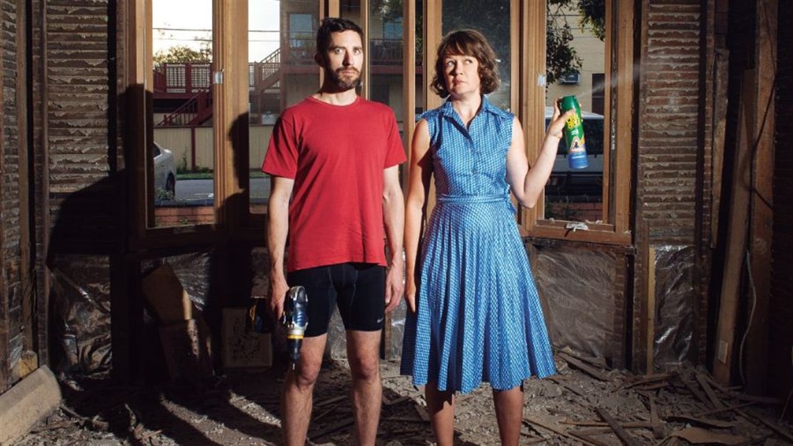 A man a woman stand in front a dilapidated picture window with a drill gun and bug repellent spray in their hands. They look puzzled at what's around them. 