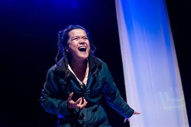 A woman with long dark hair is wearing a dark green long sleeve jacket. She leans slightly forward with her right arm bent and hand facing the ceiling, while her left hand reaches back. A white sheer curtain is to her back right.