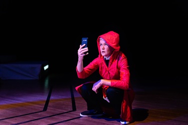 A person wearing a read hooded jumper and black pants crouches on a wooden floor. They hold up a mobile phone to their face.