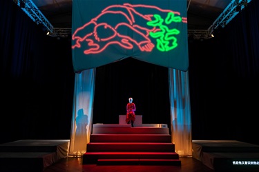 A large scale set piece is on display with coloured graffiti displayed on a large proscenium. Below the proscenium sits a person in a red outfit at the top of a set of red stairs.
