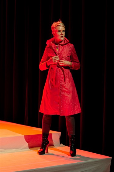 A woman with short light hair stands on a white platform wearing a red trenchcoat, black stockings and black heeled boots. A hands-free mic is taped to her right cheek. A black curtain is behind.