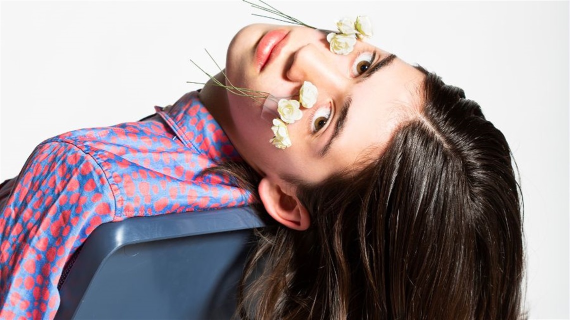 A young woman with long brown hair leans her head back off the back of a chair. Tw bunches of white flowers are taped to her cheeks with the buds towards her eyes.