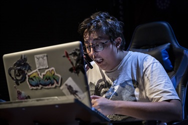 A person with short dark hair and black thin-framed glasses sits behind a computer screen. They look worried at the screen.
