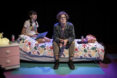 Two people sit on a bed. One person is wearing black think-framed glasses and a green jacket. The other peson has long dark hair in braided pigtails wearing a white tshirt. A pink coloured side table and blue rug complete the room.