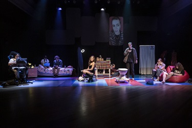 A group of young people are placed across a lit stage. They are sitting and standing in various positions. In the background is an image of a young person with light long curly hair.