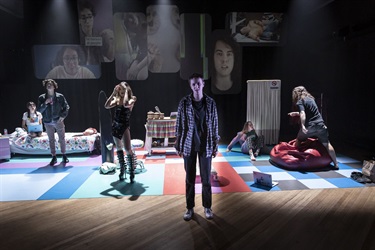 A group of young people stand in various positions across a lit stage. There are images projected on a screen in the background.