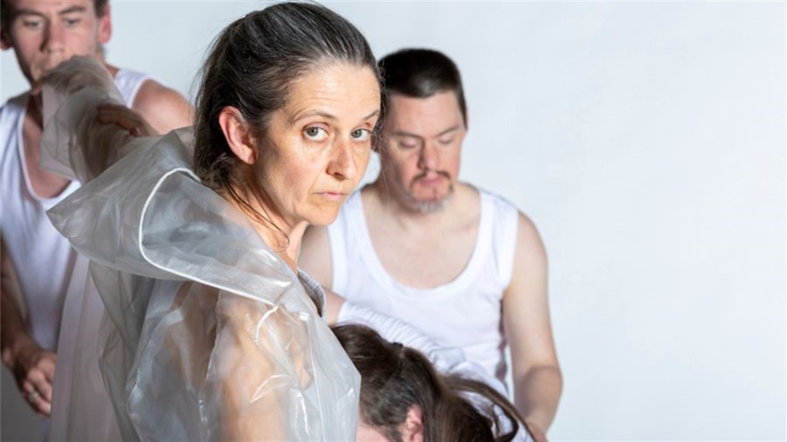 A woman with grey hair pulled back in a ponytail stands in profile with her face turned over her right shoulder. She wears a clear plastic garment. Two men are blurred in the background, both wearing white tank tops.