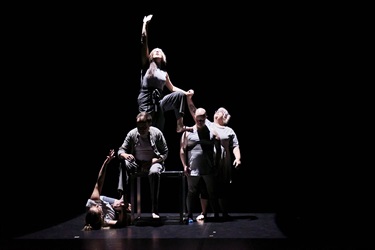 Six people gather around a table and form a human tower. The woman on top has long dark hair, with her right arm stretched tot he sky and head tilt back to look up to the sky.