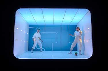 Two women stand across from on another wearing white outfits. They are contained in a white stark room lit by blue lighting.