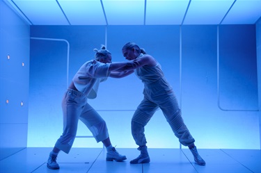 Two women wearing white outfits lean and push onto one another. They are surrounded by a stark white room lit with blue lighting.