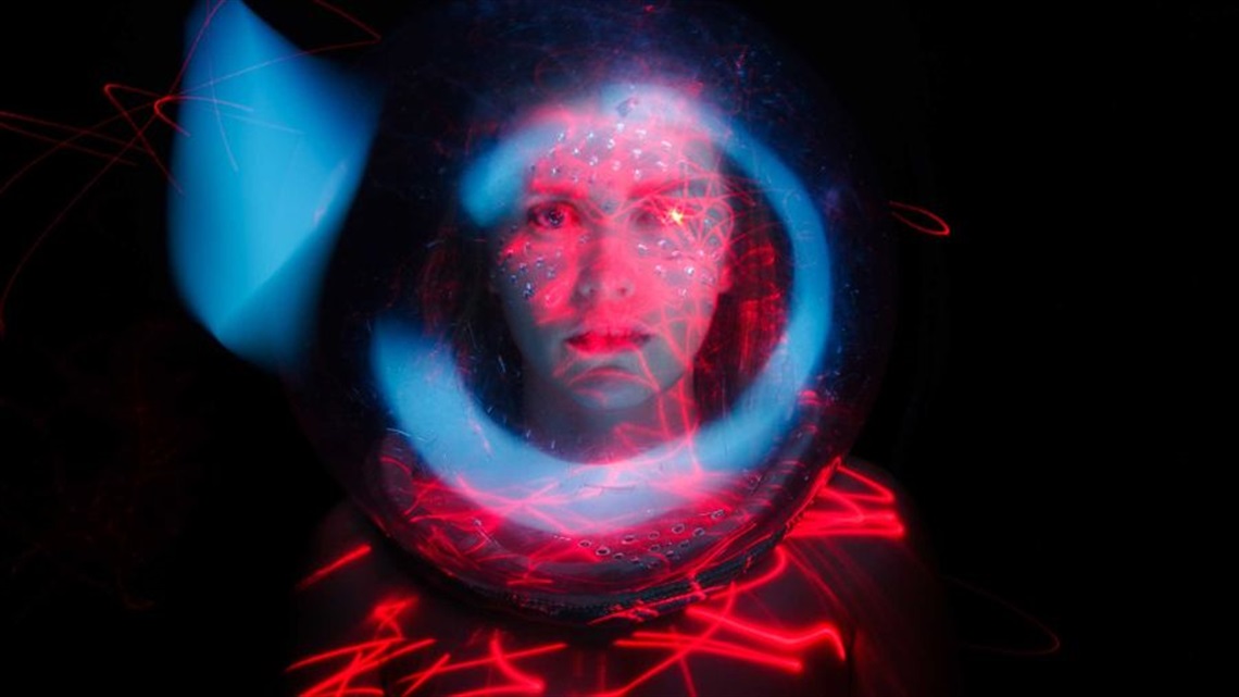 A person wearing an astronaut's outfit stars intently to the camera. Red strips of light over their upper shoulders, and a blue glow circles their face.