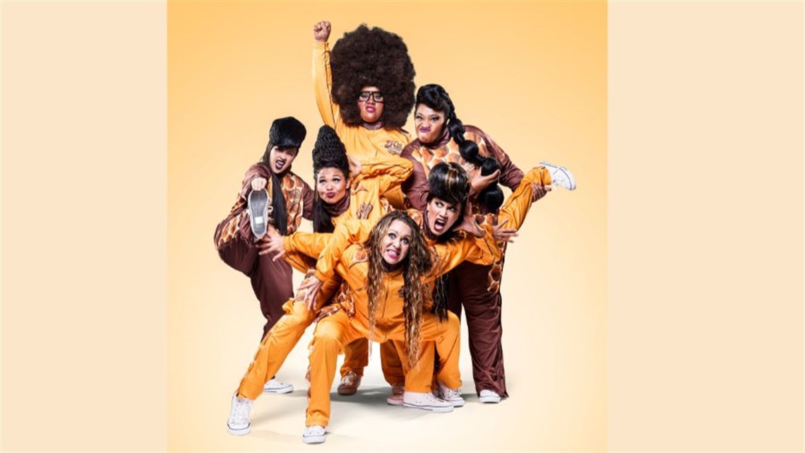 A group of Blak women cluster in a bunch wearing yellow and brown track suits. Many have large dark hair styles and are making funny faces.