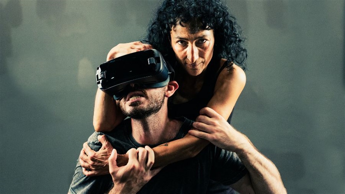 A woman with long curl dark hair stands behind a man wearing virtual reality goggles on his eyes. She wraps her arms around his neck in a loose choke hold. The man grabs her right arm with his hands.