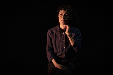 A woman wearing a dark buttoned-down shirt holds her right hand below her chin. She looks to her left to the horizon while a soft glowing light lights up the left-side of her face.