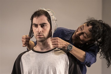 A man with dark short hair is wearing a hot on his head and a grey tshirt. He looks annoyed. A woman stands behind him leaning over his right shoulder and holds down the side of the hat on the man's head.