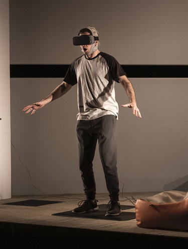 A man wearing dark trousers, a grey tshirt and virtual reality goggles on his head stands with his arms spread out to the side.