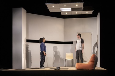 A woman with long dark curly hair and a man with short dark hair stand across from each other on a theatre set. The woman wears a dark long sleeve top and black trousers. The man wears a white tshirt and grey sweat pants.