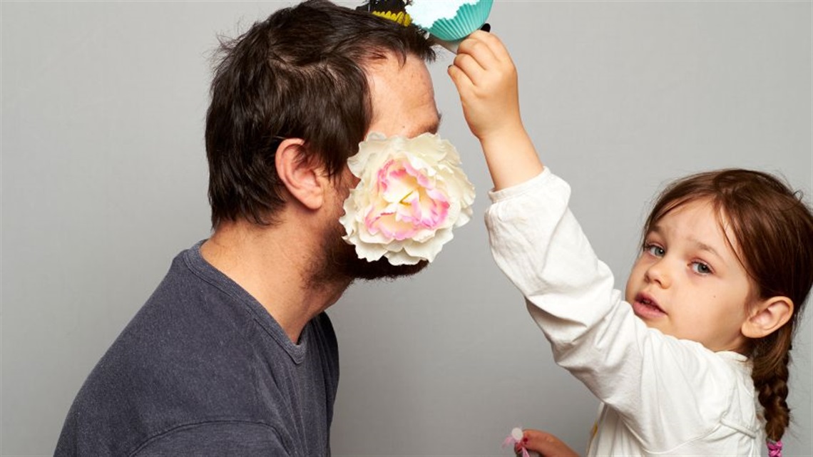 A child holds a cupcake paper to the top of the head of an adult whose face is covered by a flower.