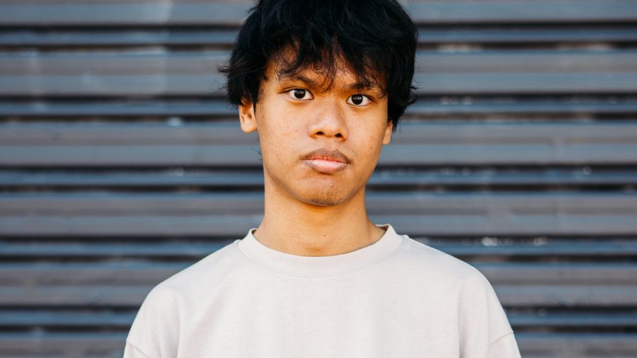 A man with short dark hair wears a light tshirt and stands in front of a grated door. He looks solemnly towards to the camera.
